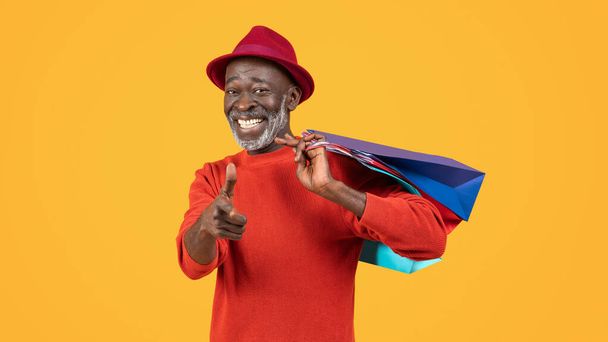 Charismatische zwarte senior met een warme glimlach, met een rode trui en hoed op, speels knipogend en wijzend terwijl hij kleurrijke boodschappentassen vasthoudt, tegen een zonnige gele achtergrond - Foto, afbeelding