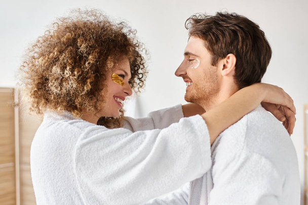 Retrato de pareja feliz con parches en los ojos abrazándose en el baño y sonriendo, mirándose - Foto, imagen