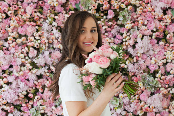 Smiling female model with rose flower in her hand standing on blooming floral blossom background. Love, 8 march, birthday and holiday woman portrait - Photo, Image