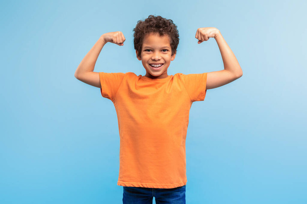 Młody czarny chłopiec w pomarańczowym t-shircie pokazuje swoje mięśnie z dużym uśmiechem, emanując pewnością siebie i siłą, na miękkim niebieskim tle - Zdjęcie, obraz