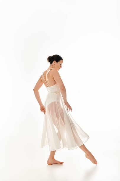 Μια νεαρή γυναίκα με ένα λευκό φόρεμα που ρέει χαριτωμένα χορεύει σε ένα σκηνικό στούντιο σε λευκό φόντο. - Φωτογραφία, εικόνα