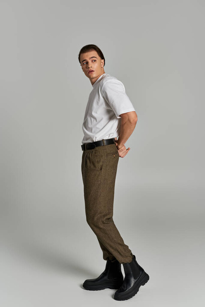 stylový mladý muž v tričku a hnědé kalhoty pózuje atraktivně na šedém pozadí a dívá se jinam - Fotografie, Obrázek