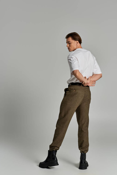 schöner junger Mann in T-Shirt und brauner Hose posiert attraktiv vor grauem Hintergrund und schaut weg - Foto, Bild