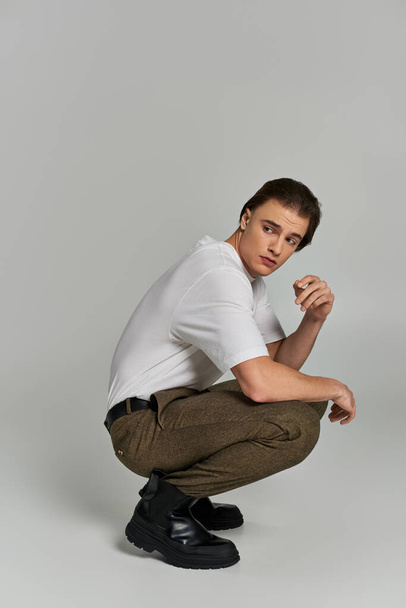 handsome elegant male model in fashionable attire squatting and looking away on gray backdrop - Foto, Imagen
