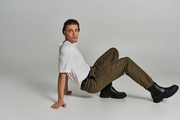 joven guapo en camiseta y pantalones marrones posando atractivamente sobre fondo gris y mirando hacia otro lado - Foto, Imagen