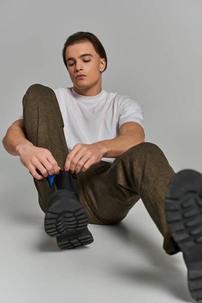 atractivo modelo masculino debonair en pantalones marrones sentado y atando sus cordones sobre fondo gris - Foto, imagen