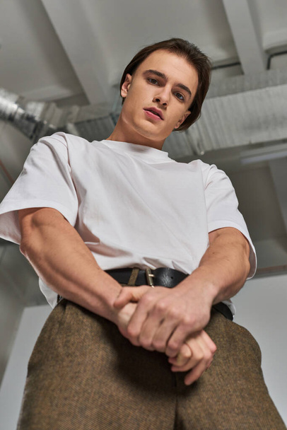 fashionable young male model in sophisticated pants looking at camera while on gray background - Φωτογραφία, εικόνα