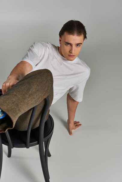 schöner stylischer Mann in Debonair-Kleidung sitzt auf einem Stuhl und schaut in die Kamera vor grauem Hintergrund - Foto, Bild
