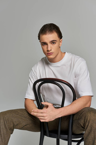 schöner trendiger Mann in Debonair-Kleidung sitzt auf einem Stuhl und blickt in die Kamera vor grauem Hintergrund - Foto, Bild