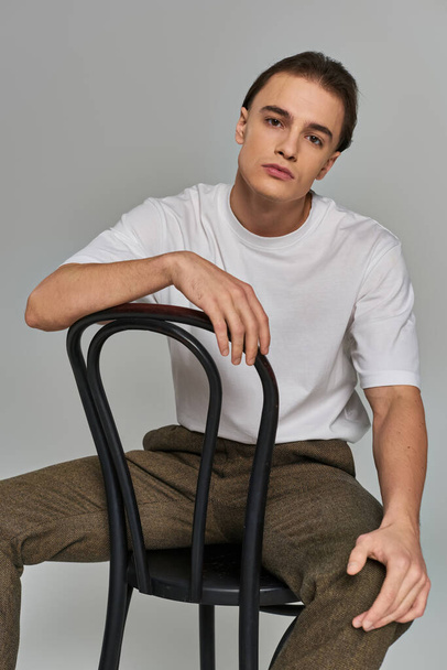 Yakışıklı, modaya uygun, güler yüzlü bir adam sandalyede oturuyor ve gri arka plandaki kameraya bakıyor. - Fotoğraf, Görsel