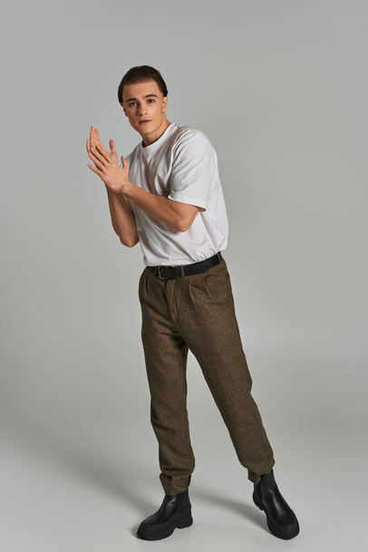 modelo masculino joven de moda en pantalones sofisticados mirando a la cámara mientras está sobre fondo gris - Foto, imagen