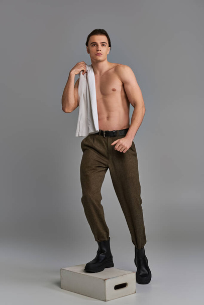 atractivo atractivo joven en pantalones marrones posando en topless sobre fondo gris y mirando hacia otro lado - Foto, Imagen