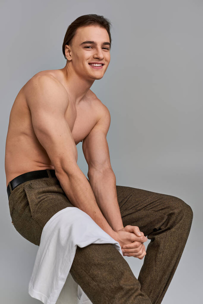 alegre seductor hombre en pantalones marrones posando en topless mientras está sentado mirando a la cámara en el fondo gris - Foto, Imagen