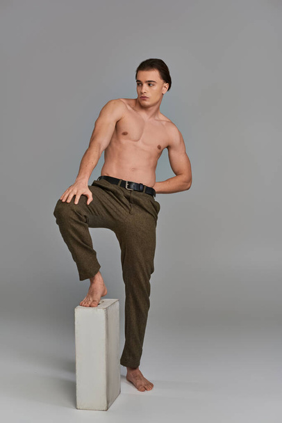 заманчивый мужчина без рубашки в коричневых элегантных брюках привлекательно позирует на сером фоне и смотрит в сторону - Фото, изображение