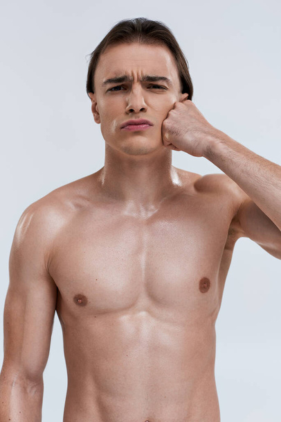 guapo atractivo emocional modelo masculino posando en topless con el puño cerca de la mejilla y mirando a la cámara - Foto, imagen