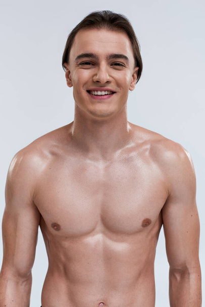 alegre seductor joven modelo masculino posando en topless y sonriendo felizmente a la cámara en el fondo gris - Foto, imagen