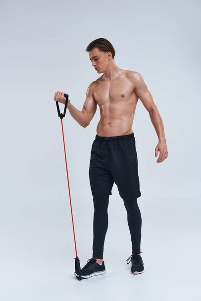 seductor sin camisa joven deportivo en pantalones negros de entrenamiento con expansor de fitness y mirando hacia otro lado - Foto, imagen