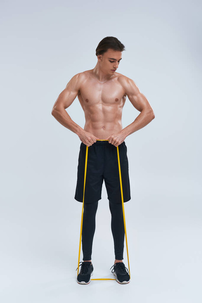 alluring athletic young man in black pants posing topless with resistance band and looking away - Photo, Image