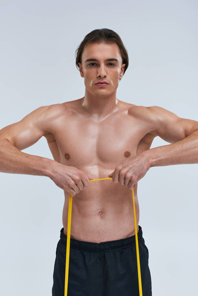 sporty alluring young man posing topless while training with resistance band and looking at camera - Photo, Image