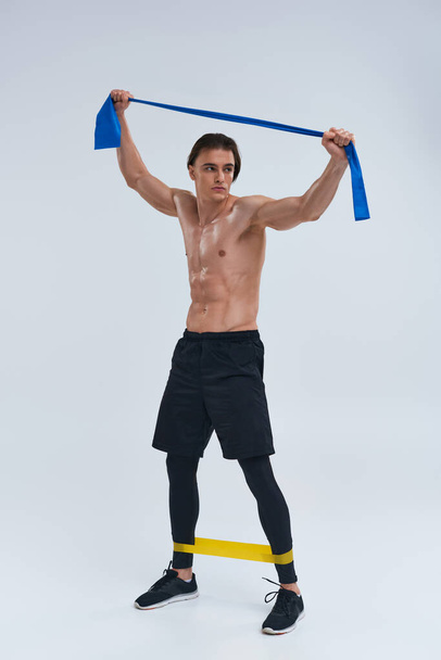 alluring sporty young man in black pants posing topless with resistance bands and looking away - Photo, Image