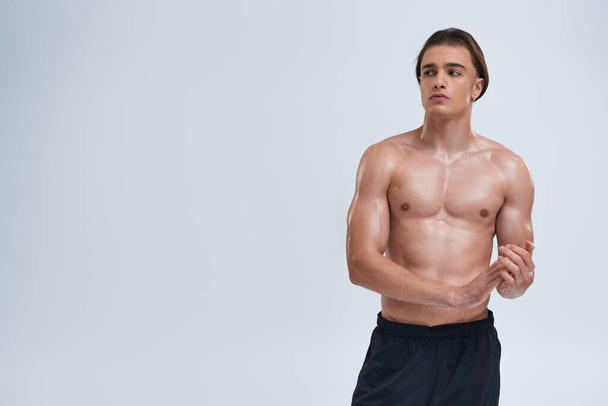 athletic good looking young man posing shirtless in black pants and looking away on gray background - Photo, Image