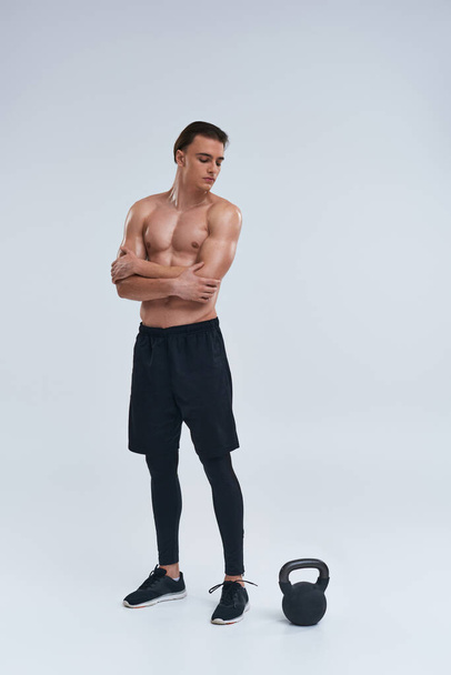 gut aussehender sportlicher Mann in schwarzer bequemer Hose posiert auf grauem Hintergrund und schaut auf die Kettlebell - Foto, Bild