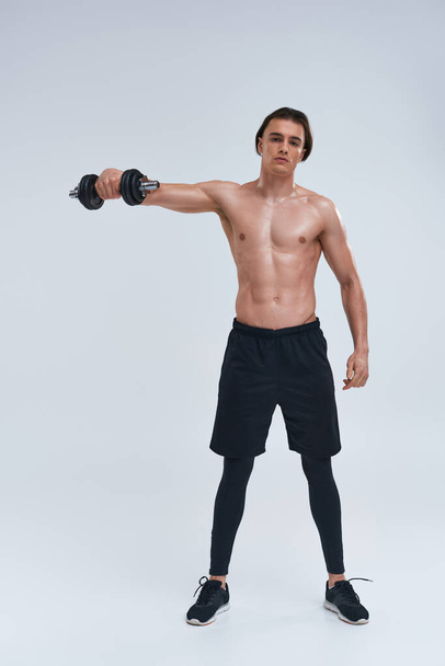 attractive athletic man posing topless exercising actively with dumbbell and looking at camera - Photo, Image