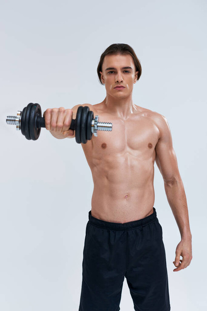 attractive athletic man posing topless exercising actively with dumbbell and looking at camera - Photo, Image