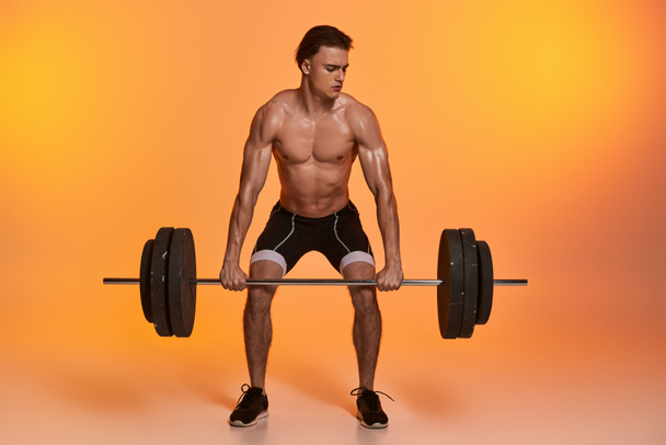 seductor hombre musculoso sin camisa en pantalones negros haciendo ejercicio con barra de pesas sobre fondo naranja vibrante - Foto, Imagen