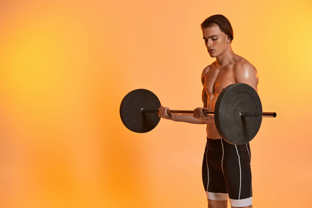 活気に満ちたオレンジ色の背景でベルで運動する黒パンツのハンサムシャツレス筋肉男性 - 写真・画像