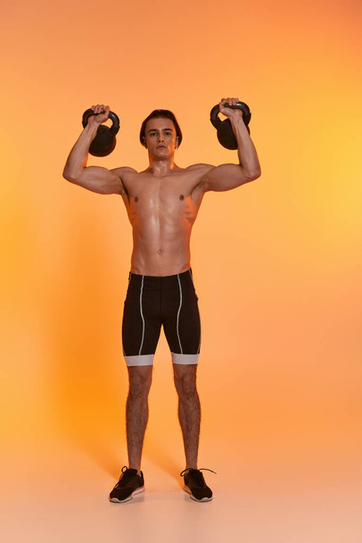 appealing sporty shirtless man exercising with kettlebells and looking at camera on vivid backdrop - Photo, Image