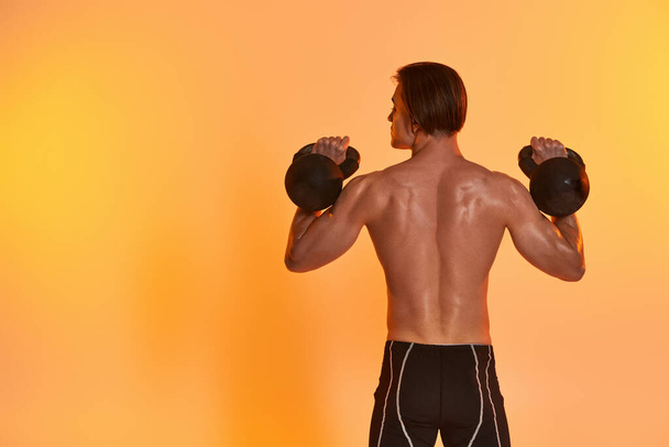 back view of young muscular man posing topless while exercising with kettlebells on orange backdrop - Photo, Image