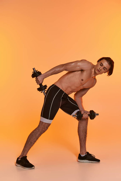 hombre musculoso sin camisa de buen aspecto en pantalones cortos deportivos negros entrenando con mancuernas en un fondo vívido - Foto, Imagen