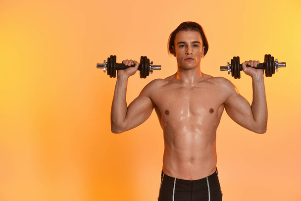 привлекательный спортивный молодой человек позирует без верхней одежды, позируя с гантелями и глядя в камеру - Фото, изображение