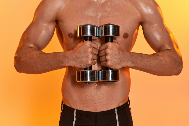 recortado vista de sexy musculoso hombre posando en topless durante el entrenamiento con mancuernas en naranja telón de fondo - Foto, imagen