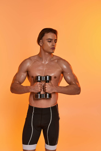 appealing muscular shirtless man in black sport shorts training with dumbbells on vivid backdrop - Photo, Image