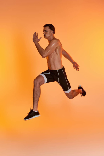 atrakcyjny mężczyzna bez koszulki w czarnych spodenkach ćwiczenia aktywnie i patrząc daleko na pomarańczowym tle - Zdjęcie, obraz