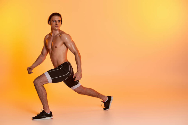 appealing shirtless man in black shorts exercising actively and looking away on orange backdrop - Photo, Image