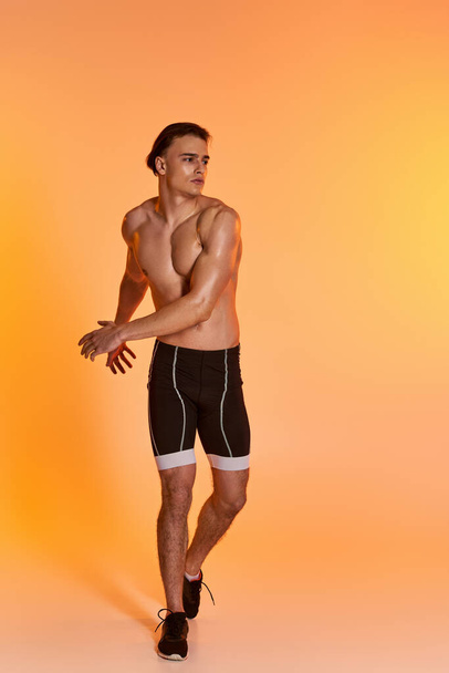 hombre guapo sin camisa en pantalones cortos negros haciendo ejercicio activamente y mirando hacia otro lado en el fondo naranja - Foto, imagen