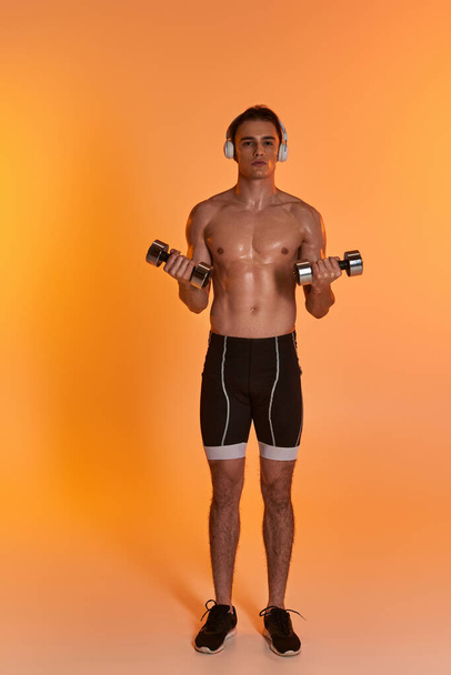 atractivo hombre musculoso en pantalones cortos con auriculares haciendo ejercicio con pesas y mirando a la cámara - Foto, Imagen