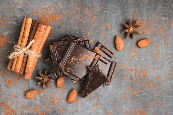 Heap of delicious dark chocolate pieces or cubes, chopped, broken chocolate bar, almond nut, cinnamon sticks and star anise on a dark background, top view. - Photo, Image