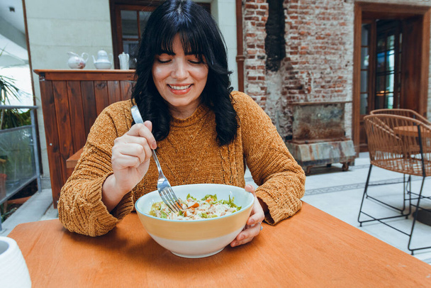 front view of young Latin woman with long black hair, wearing brown sweater, is happy sitting in restaurant watching bowl of delicious salad she is going to eat, restaurant concept, copy space - Photo, Image