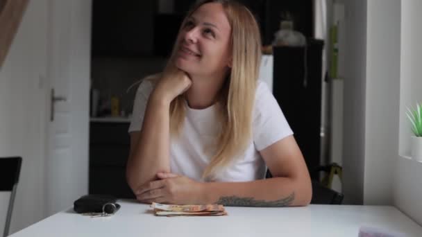 a girl with blond hair sits indoors at a table, with her hands folded, and the table looks thoughtfully to the side, on the table are euros in the amount of 50 salaries. High quality FullHD footage - Footage, Video