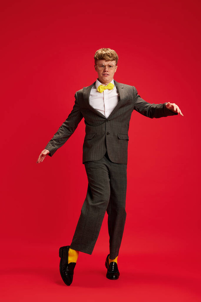 Full-length portrait of young man in formal wear with yellow bright socks dancing against red studio background. Concept of business, youth, human emotions, lifestyle - 写真・画像