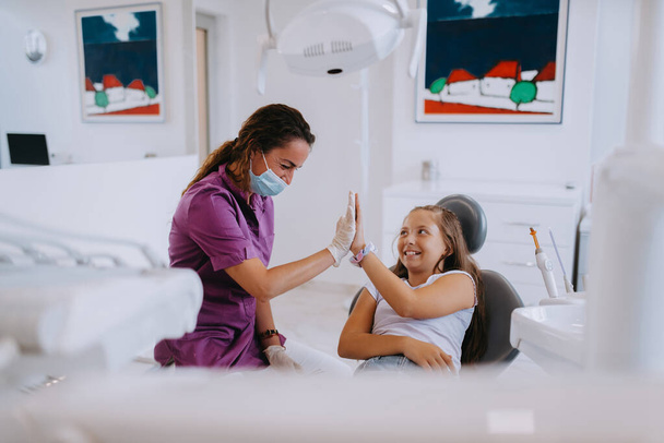 Ein fürsorglicher Zahnarzt führt ein fröhliches Gespräch und unterhält ein kleines Mädchen nach einer Zahnbehandlung, wodurch eine kindgerechte und angenehme Atmosphäre in der Zahnarztpraxis entsteht - Foto, Bild