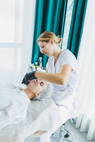 皮膚科医は,マッサージテーブルに横たわっている女性の顔を扱います. フェイシャルスキンケア。 女性の美容師は女性の顔にマスクを適用します - 写真・画像