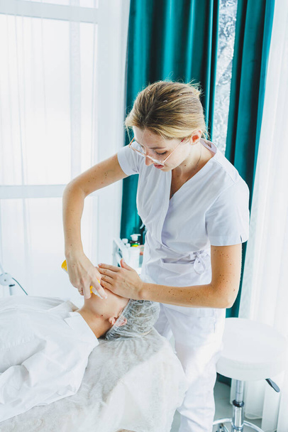 Een dermatoloog behandelt het gezicht van een vrouw die op een massagetafel ligt. Gezichtsverzorging. Een vrouwelijke schoonheidsspecialiste brengt een masker aan op het gezicht van een vrouw - Foto, afbeelding