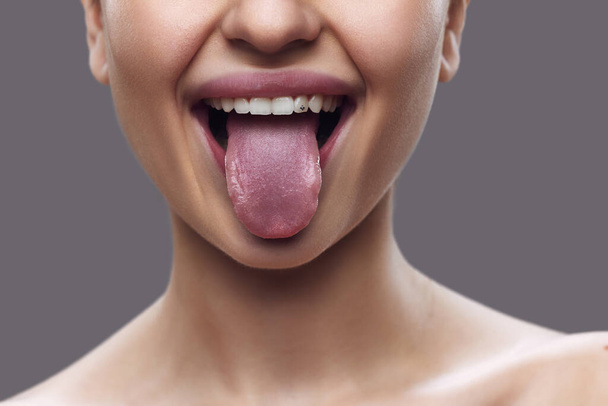 Vystřižená fotka. Detailní záběr ženských úst s jazykem, zvýrazněný jemným leskem na rtech, na šedém pozadí. Koncepce technik pro zlepšení kloubové a svalové kontroly v řeči. - Fotografie, Obrázek
