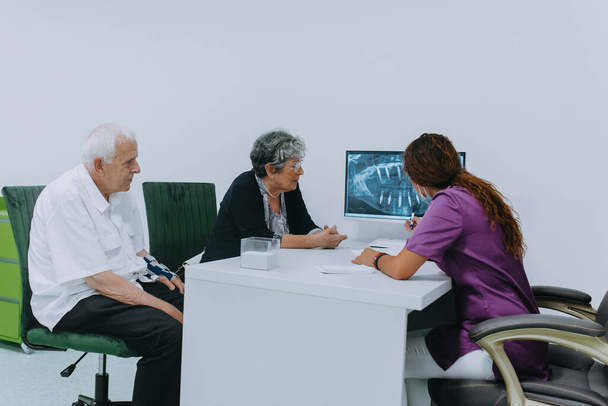 Um casal de idosos se envolve em uma discussão pensativa com seu dentista sobre as opções modernas de prótese dentária em um ambiente de consultório odontológico contemporâneo.. - Foto, Imagem