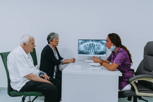 Una pareja de ancianos se involucra en una discusión reflexiva con su dentista sobre las opciones de prótesis dentales modernas en un entorno de consultorio dental contemporáneo.  - Foto, imagen